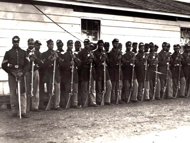A group of approximately twenty Union soldiers, appearing to be African American, standing in a line in front of a white wooden building. Each soldier is dressed in Union Army uniforms and holding a rifle upright. The black-and-white photograph dates to the Civil War era.